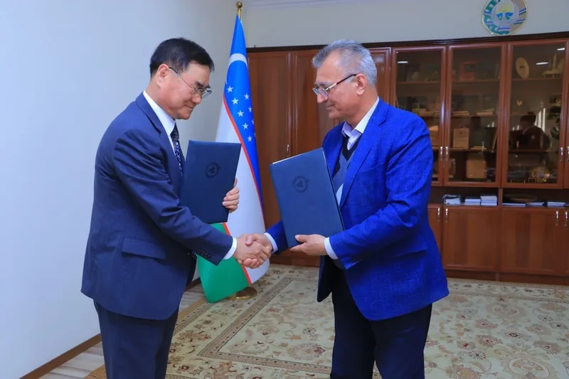 Two participants shake hands while holding blue document folders in front of the Uzbekistan flag